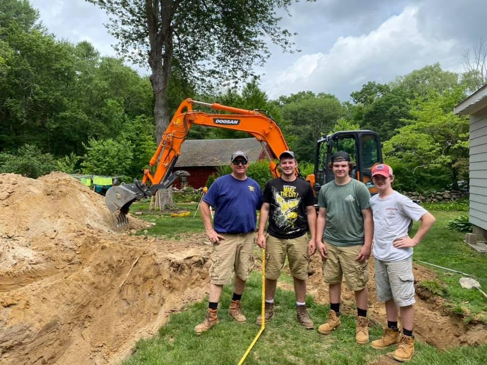 Four Parker family and crew members standing together at an excavation site in front of an orange excavator